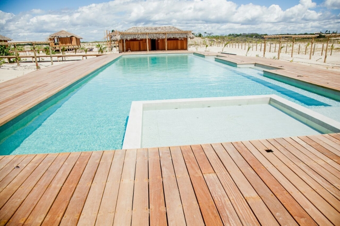 A imagem mostra uma grande piscina, cercada por um piso de madeira e em meio a areia branca de praia
