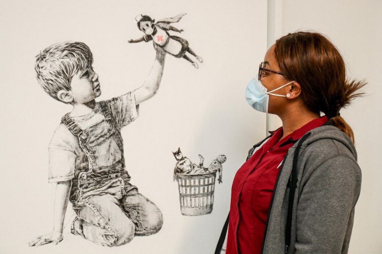 Profissional de Saúde do University Hospital Southampton diante de de obra de Banksy chamada "Game Changer", na qual criança brinca com boneca de super-heroi que é uma enfermeira