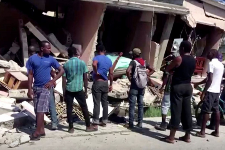 People stand in front of a collapsed building following an earthquake, in Les Cayes, Haiti, in this still image taken from a video obtained by Reuters on August 14, 2021.   REUTERS TV via REUTERS THIS IMAGE HAS BEEN SUPPLIED BY A THIRD PARTY. MANDATORY CREDIT.