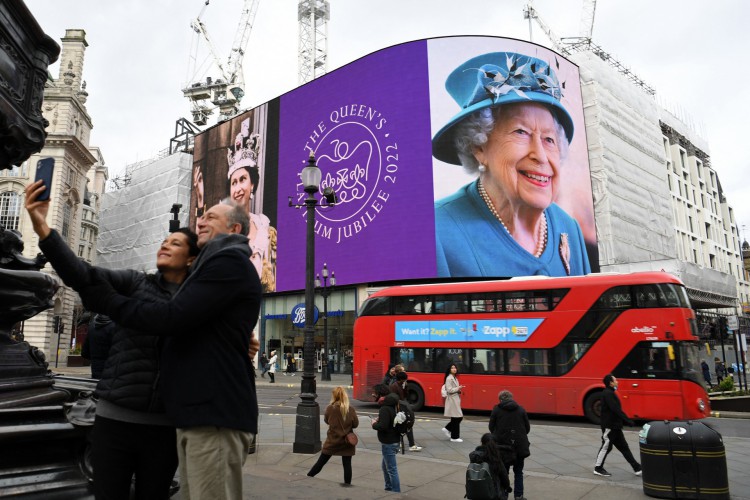 Um casal tira uma selfie enquanto um ônibus vermelho de Londres passa, enquanto imagens da rainha britânica Elizabeth II são exibidas nas grandes telas digitais em Piccadilly Circus, no centro de Londres, em 6 de fevereiro de 2022, para marcar o início do Ano do Jubileu de Platina de Sua Majestade. A rainha Elizabeth II se torna no domingo a primeira monarca britânica a reinar em sete décadas