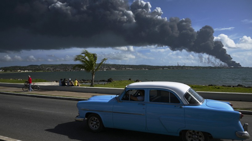 Fumaça preta de um tanque de óleo em chamas é vista enquanto um velho carro americano passa po...
