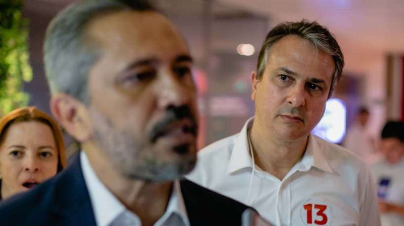 Elmano, imagem desfocada, com Camilo Santana ao fundo, em evidência, durante entrevista coletiva 