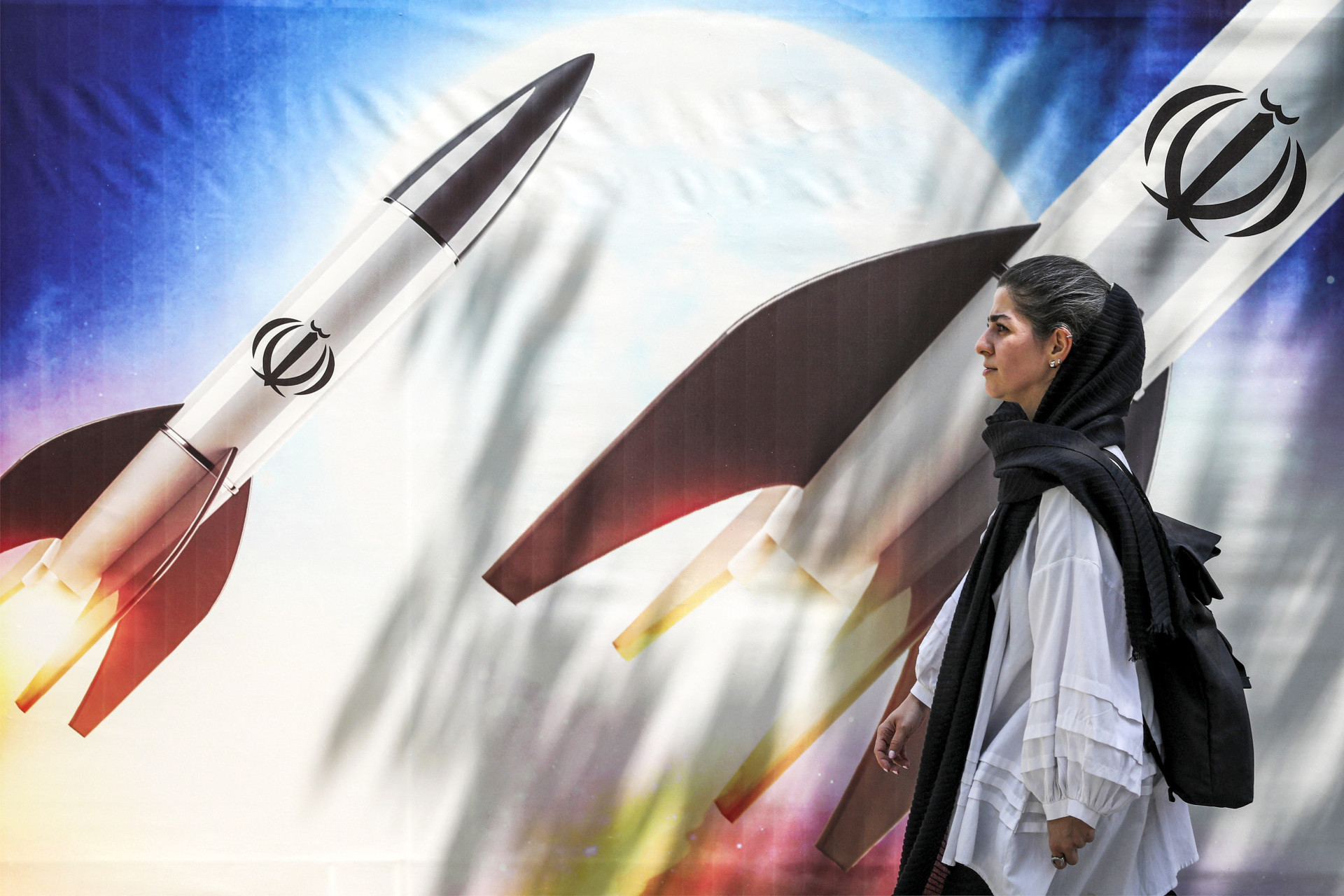 ￼MULHER passa por banner que representa lançamento de mísseis com o emblema do Irã (Foto: ATTA KENARE / AFP)