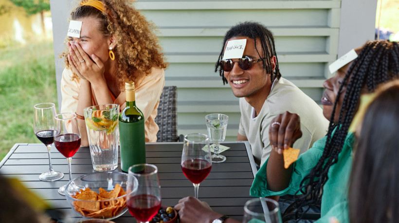 Group of young people laughing and playing Guess who game while sitting at table outdoors in Summer
