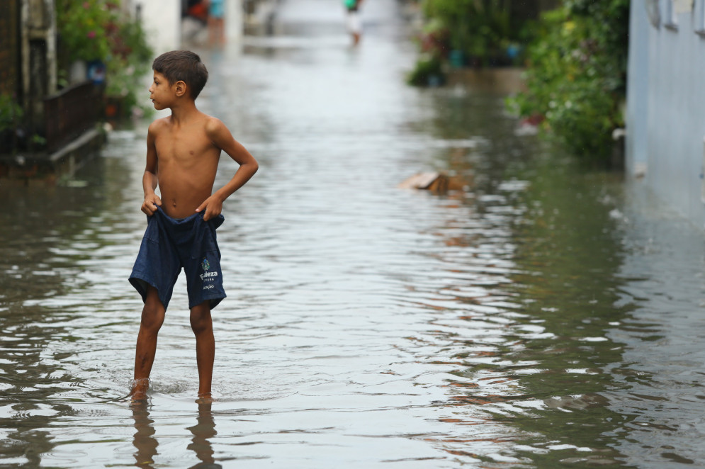 Imagem da chuva em Fortaleza. Menino enfrenta as águas em rua alagada vestindo apenas um shorts de fardamento escolar