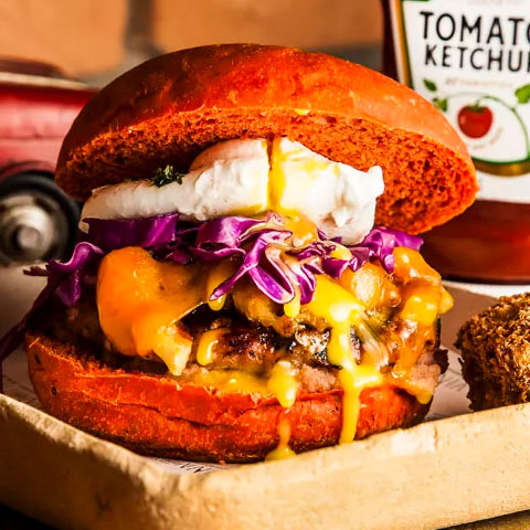 O hambúrguer Porco Feliz contém costela suína e panceta, levando queijo cheddar, cebola caramelizada e purê de maçã. A cor do pão é diferente, puxada para um vermelho alaranjado.