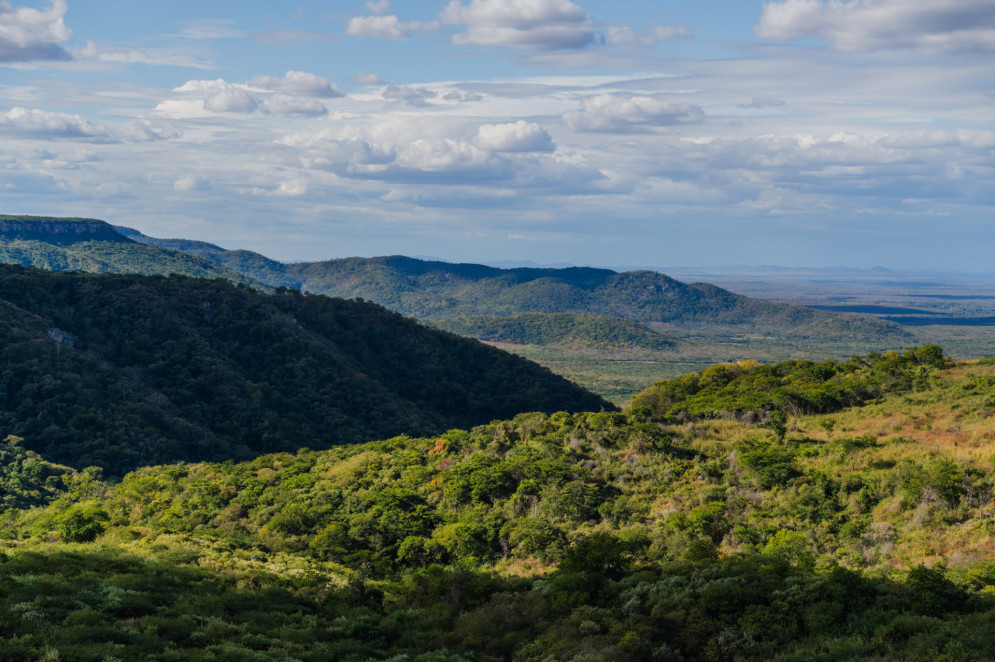 A imagem mostra uma paisagem serrana coberta por vegetação verde, com colinas que se estendem até o horizonte. O céu está parcialmente nublado, compondo um cenário natural amplo e sereno