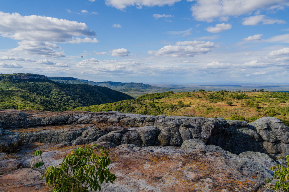 A imagem mostra uma paisagem natural com formações rochosas em primeiro plano e montanhas cobertas por vegetação ao fundo. O céu está azul com nuvens brancas espalhadas