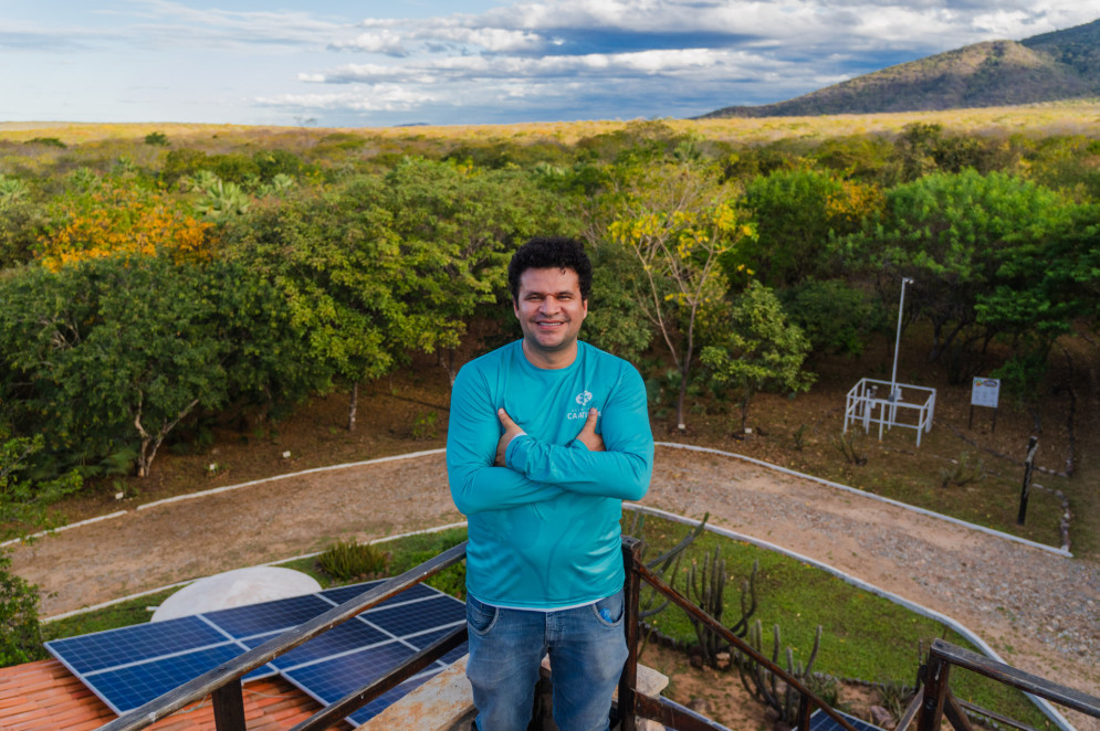 A imagem mostra um Gilson sorridente de braços cruzados vestindo camiseta azul, posando em um mirante com painéis solares ao fundo. Ele está cercado por vegetação nativa e montanhas ao longe sob um céu parcialmente nublado