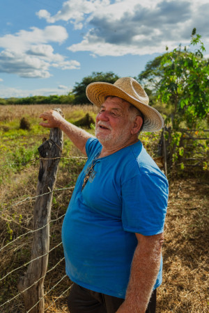 Homem idoso de camisa azul e chapéu de palha aponta para uma plantação atrás de uma cerca de madeira e arame