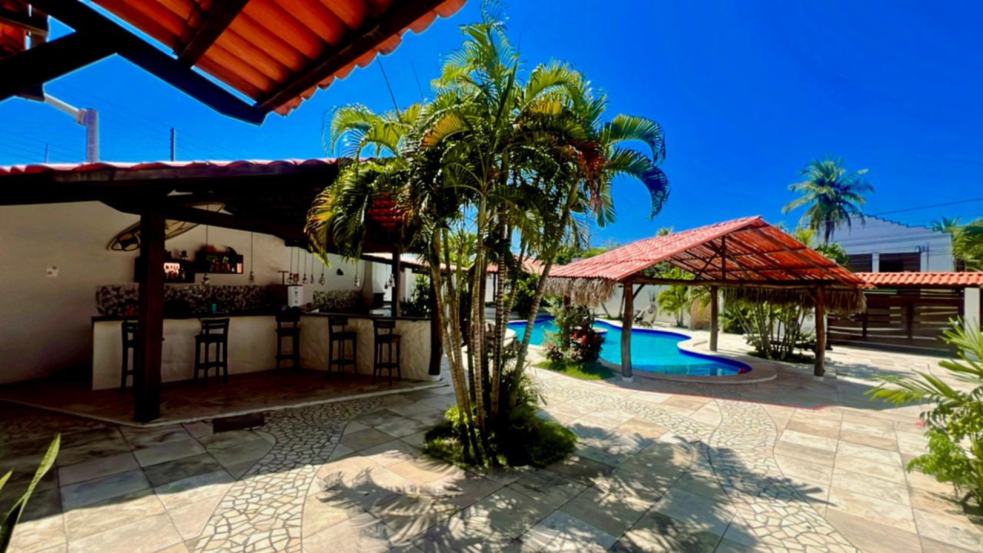 A estrutura inclui piscina, área de lazer com churrasqueira e bar, suítes equipadas com ar-condicionado, chuveiro aquecido por energia solar, escrivaninha, TV Smart, frigobar, e camas confortáveis