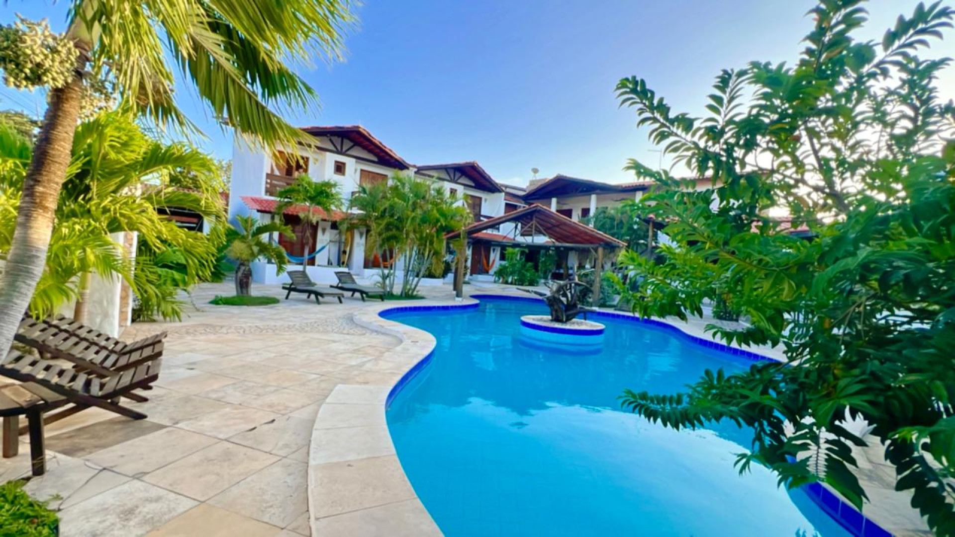 A estrutura inclui piscina, área de lazer com churrasqueira e bar, suítes equipadas com ar-condicionado, chuveiro aquecido por energia solar, escrivaninha, TV Smart, frigobar, e camas confortáveis