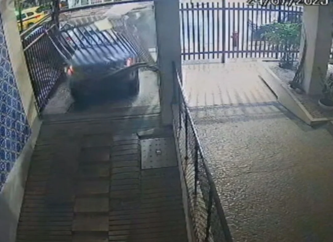 O carro avança atingindo o portão do estacionamento do edifício. O impacto é tão grande que o portão é arrancado.