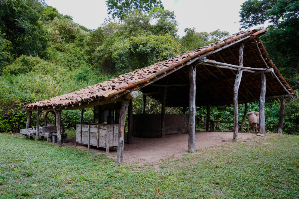 A imagem mostra uma Casa de Farinha rústica com estrutura de madeira e telhado de barro, localizada em meio à vegetação. No interior e arredores, há utensílios tradicionais usados na produção de farinha de mandioca