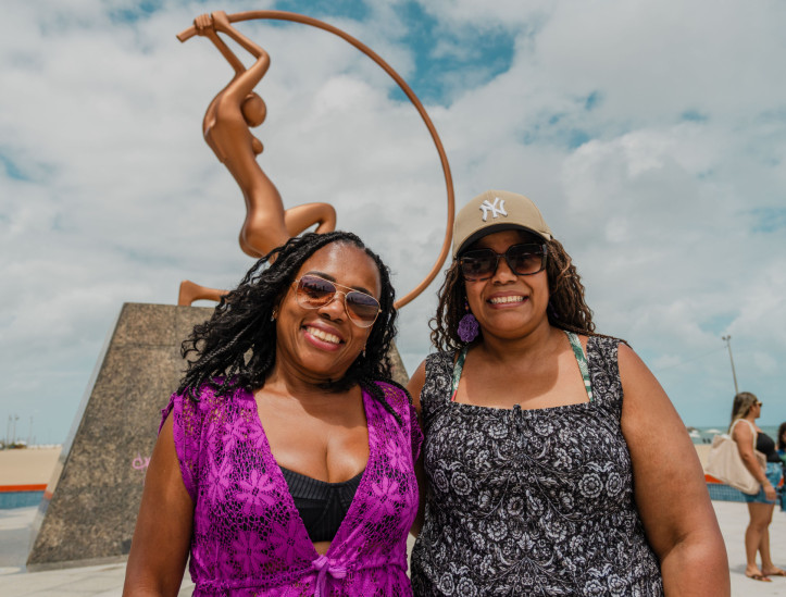 FORTALEZA, CEARÁ, BRASIL, 23-11-2025: Reportagem sobre as condições estruturais de estátuas e esculturas da orla da Beira Mar e Praia de Iracema. Na foto, Nilza Valquíria de Oliveira, 52, turista natural de Belo Horizonte, e ao fundo a estátua "Iracema Guardiã", do artista Zenon da Cunha Mendes Barreto, de 1995, na Praia de Iracema. (Foto: Fernanda Barros/ O Povo)