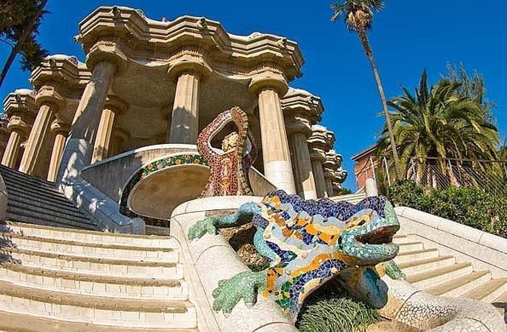 A entrada principal do parque é marcada por duas casas de contos de fadas, com telhados ondulados e mosaicos coloridos. Essas construções lembram doces cobertos de açúcar, reforçando o caráter lúdico do espaço. O portão conduz os visitantes a uma escadaria monumental. No centro, destaca-se a famosa escultura do dragão de cerâmica. É o primeiro contato com o universo mágico de Gaudí.