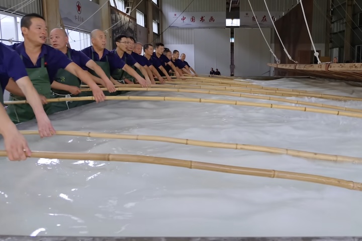 A fabricação das folhas ocorre em uma sala de mais de 11 metros de comprimento, onde a polpa é distribuída manualmente na água, com a ajuda de varas de bambu, até formar uma superfície uniforme.