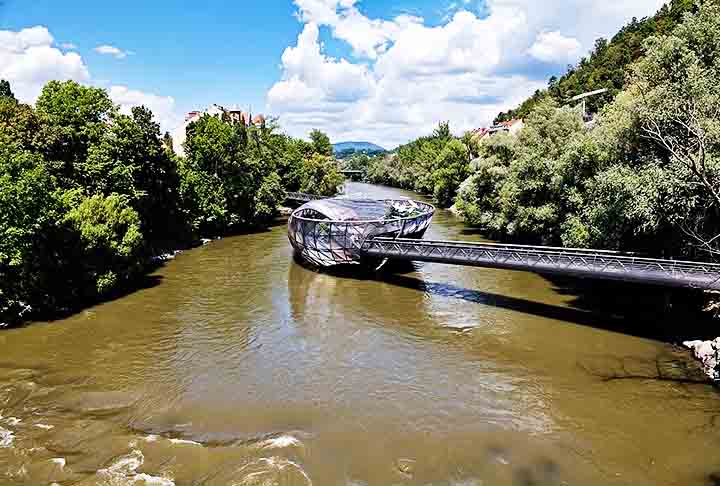 Inaugurada em 2003, a Ilha flutuante artificial Murinsel é composta por uma plataforma em forma de concha que abriga um café, um espaço para eventos e uma área de recreação, conectando as margens do rio com passarelas.