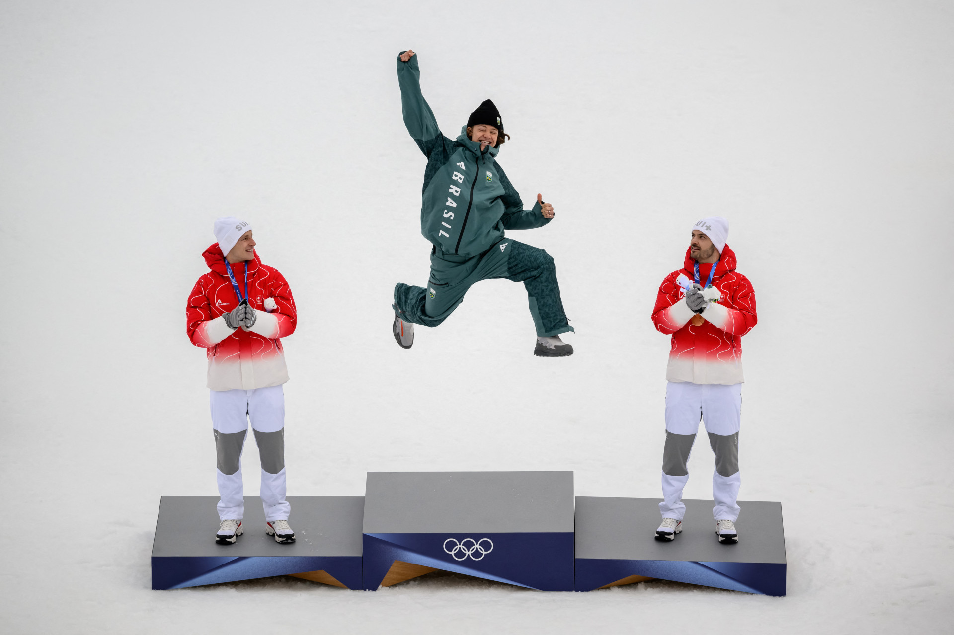 ￼O MEDALHISTA Lucas Pinheiro Braathen salta no pódio, ladeado pelos suíços Marco Odermatt (esquerda) e Loic Meillard (Foto: Fabrice COFFRINI / AFP)