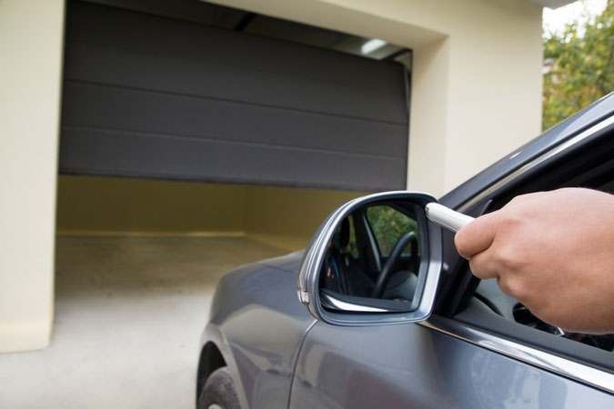 A imagem mostra um carro prestes a entrar em uma garagem. A garagem está com o portão semi-aberto, indicando que está em processo de abertura. Do carro, que é cinza, podemos ver uma mão saindo pela janela, segurando o controle do portão, para abri-lo. 
